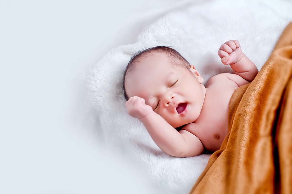 A baby lies on a white furry surface and underneath an orange blanket.